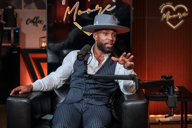 Man wearing a wide-brim gray hat, white shirt, and pinstripe vest and pants speaking into a podcast microphone while seated in a studio chair, with a neon “Moore” sign glowing behind him.