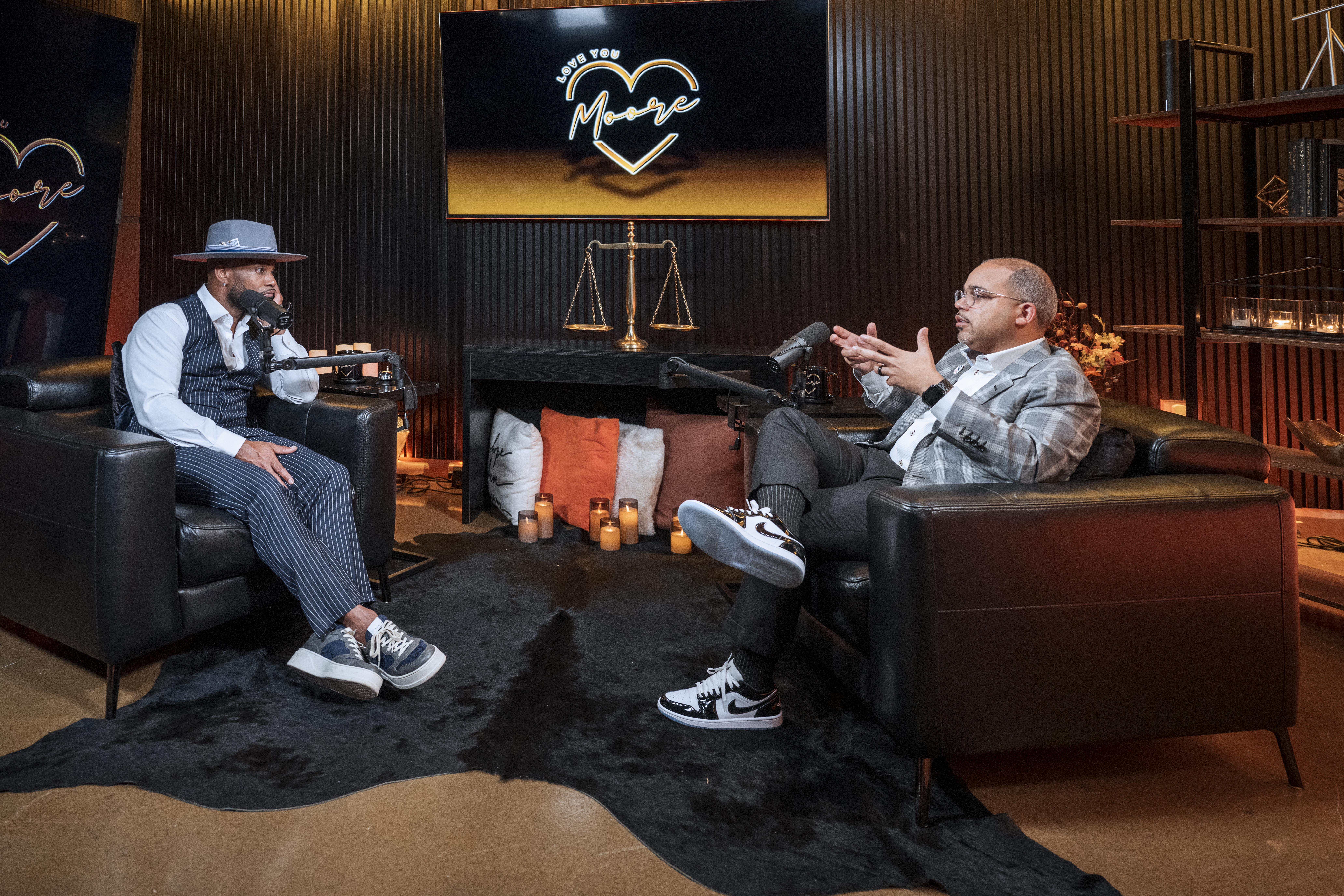 Two men seated in leather chairs during a podcast conversation, speaking into microphones in a warmly lit studio with a neon “Love You Moore” sign and decorative scales displayed behind them.