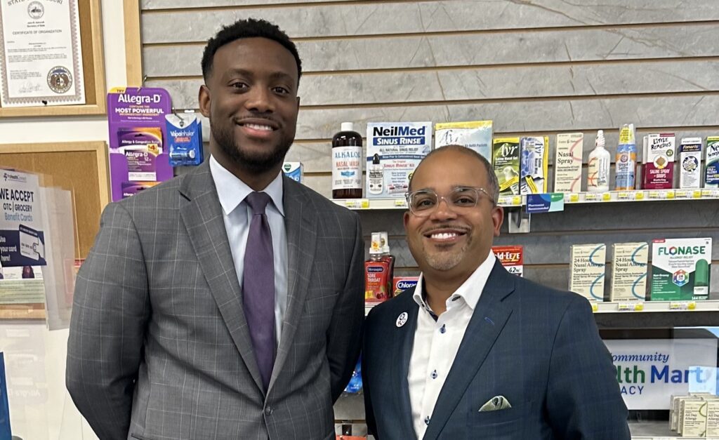 Roy Whitley, President and CEO of Rx Outreach, and Marcus Howard, CEO of GreaterHealth Pharmacy & Wellness, stand together smiling inside a community pharmacy. Roy wears a navy blazer and glasses, while Marcus wears a gray suit and tie. Pharmacy shelves stocked with over-the-counter health products are visible in the background, representing the partnership between Rx Outreach and GreaterHealth to expand medication access and pharmacy services in the St. Louis community.